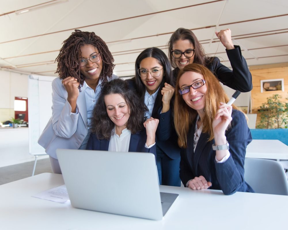 Happy businesswomen celebrating success. Cheerful multiethnic female colleagues raising fists and using laptop in office. Winner gesture concept
