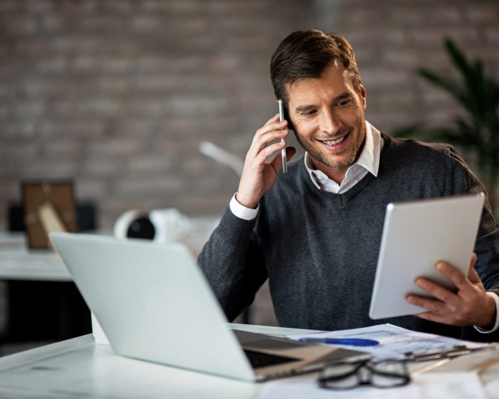 Happy entrepreneur communicating over mobile phone while working on touchpad in the office.